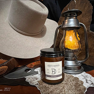 Dirty Hands candle displayed with cowboy hat, lantern, leather belt, and cowhide, this link will open in a new tab.