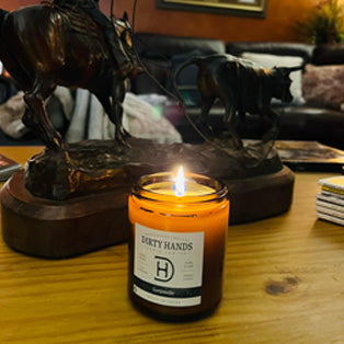 Lit Dirty Hands lavender candle on table with western-style sculpture in the background, this link will open in a new tab.