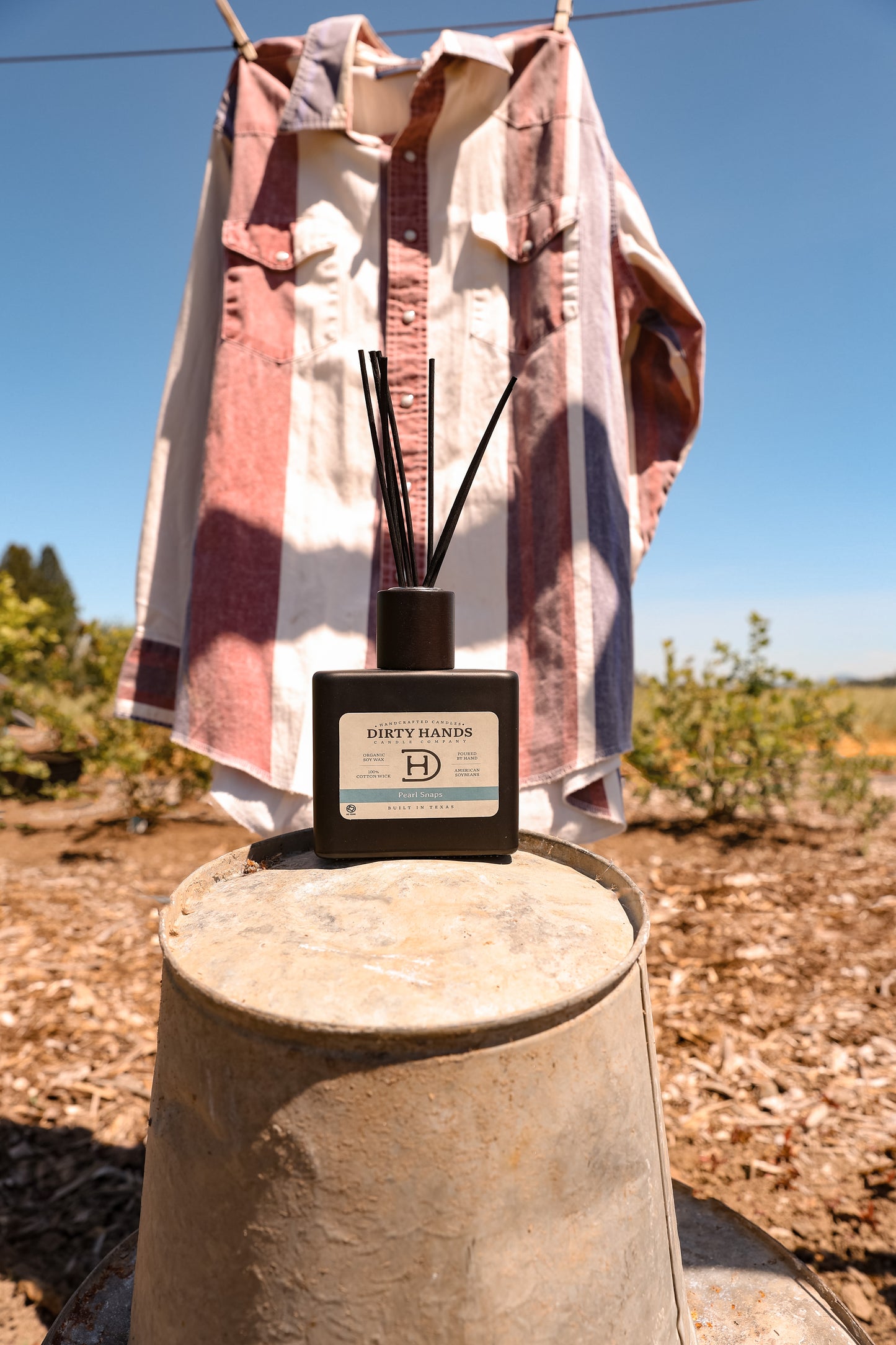 Pearl Snaps Reed Diffuser sitting on a bucket with a Pearl Snap Shirt in the background